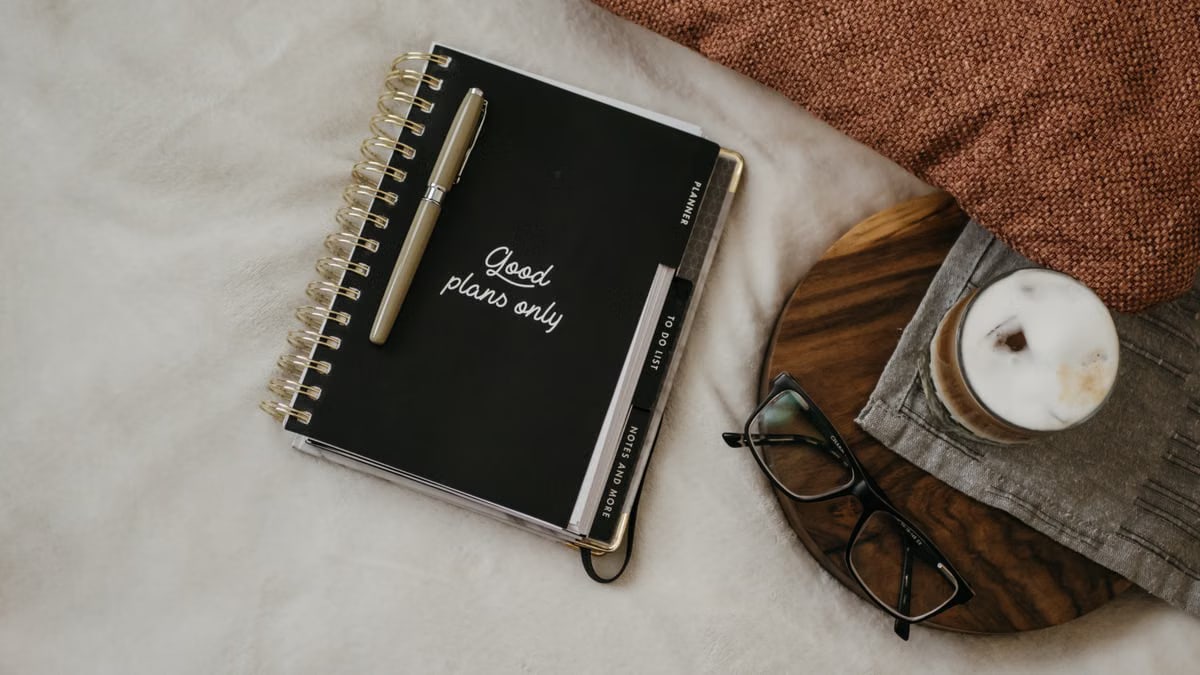 High-angle view of a notebook with 'Good plans only' sign, glasses, and coffee on a cozy setup.